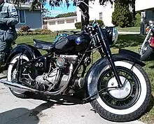 A mildly customized Sunbeam S7 motorcycle sits along the edge of a driveway.  It shows evidence of normal wear and tear, but appears to be in very good condition.  It's all black, with a vaguely egg-plant shaped fuel tank that features rubber pads for the rider's knees, and white-wall tires.