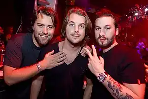 Three middle-aged men, all wearing black shirts, pose for a photograph during a DJ set