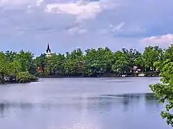 A landscape image; in the foreground can be seen the waters of the Svět pond, with light ripples. Behind them can be seen thick and vibrantly green foliage, out of which rises a steeple from the town of Třeboň. The sky is overcast.