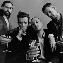 In a black-and-white image, three of the four band members of The 1975 are seen holding wine glasses while the lead singer, Matty Healy (center left) smokes a cigar.