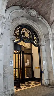 Detail of a doorway leading to the Ansonia. The doorway is arched and contains elaborate ironwork. There is a revolving door just inside the arched doorway.