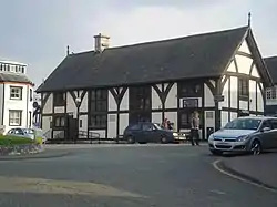 A half timbered building of two floors, with four sets of leaded windows to the front aspect and one set to the side. The build has a steep, slate roof, with a single chimney placed left of centre. Steps and a ramp lead up to its single visible entrance