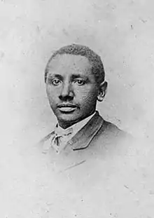 Head of a young black man with close-cropped hair wearing a dark suit coat over a shirt and tie.