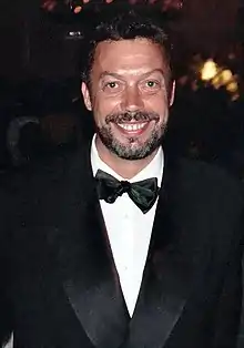 A bearded man, wearing a white shirt, black bowtie, and black jacket, smiles at the camera.