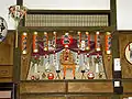 A large, decorative alcove shrine, with red paper lanterns decorated with black text strung across the top, a miniature torii gate in front and two red daruma dolls at either side.