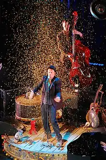 A man performing on-stage, surrounded by confetti. Several instruments surround him and behind him a man is playing a double bass.