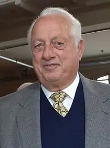 A man in a gray suit jacket, navy sweater, white shirt, and gold tie