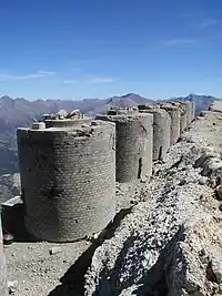 A row of round brick turrets can be seen in the foreground. A mountain range occupies the background.