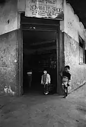 A black and white photo of two children in front of a store with a sign that says: "NON-WHITE SHOP This notice is displayed in accordance with the provisions of the Shop Hours Ordinance. 1959."