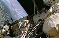 A man dressed in a white spacesuit with a red stripe seen clinging to the end of a boom-like crane, moving over a white space station module. Various trusses, solar arrays and other structures project from the module, and the Earth is visible in the background.