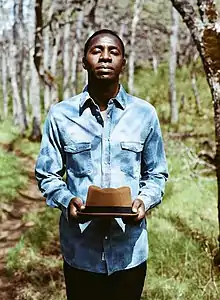 Black male standing outdoors, wearing light blue denim shirt and holding a brimmed hat, looking directly at camera