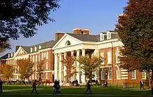 Exterior of a three-story building in red bricks, white trim with ornate columns, and students walking about a green area with trees