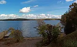 A view across a body of water with some docks on the near shore at the bottom. Woodlands with trees showing some autumn color are visible across it, and along the shoreline at the left of the image.