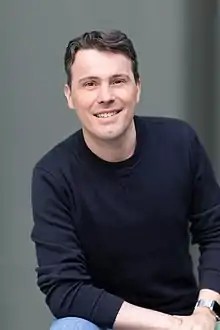 "Vincent Pilette looking toward the camera wearing a dark color long-sleeve shirt. A wristwatch and blue jeans are visible. The background is neutral gray."