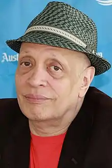 Head and shoulders of man with drooping eyelids wearing black fedora, black shirt without a collar, black jacket, and mostly grey short trimmed beard.