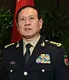 a smiling man with his mouth open and looking rightwards, wearing a light green military uniform with insignia and badges