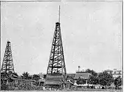 Photograph of Westinghouse "Old No.1" natural gas derrick in daytime, sometime between June 1884 and end of 1889, located at the Solitude estate in present-day Westinghouse Park.  The Westinghouse carriage house/laboratory and the mansion are seen on the right.