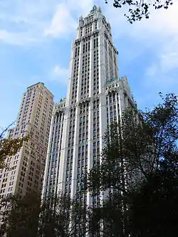 Color photo of a skyscraper with trees in the foreground and a tall but significantly shorter building to the left